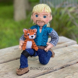 May include: Crocheted doll of a man with blonde hair and a mustache, wearing a blue shirt, jeans, and brown boots. He is holding a crocheted tiger cub. The doll is sitting on a wooden surface. The text "@craftyiscool" is visible on the surface.