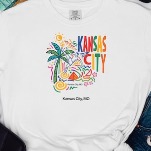 Peut inclure: Un t-shirt blanc &agrave; manches courtes avec un imprim&eacute; graphique color&eacute; portant le texte Kansas City. Le motif comprend un palmier, un soleil, un joueur de football et des formes abstraites en bleu, vert, jaune, orange et rose. Le texte Kansas City, MO est imprim&eacute; sous le motif principal.