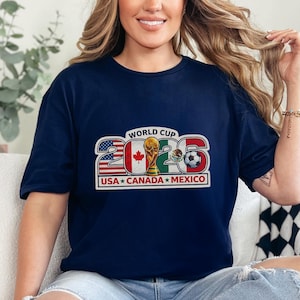 May include: Navy blue t-shirt with a graphic design for the 2026 World Cup, featuring the flags of the USA, Canada, and Mexico, a soccer ball, and a trophy. The text "WORLD CUP 2026" is displayed above the flags. The shirt is worn by a person.
