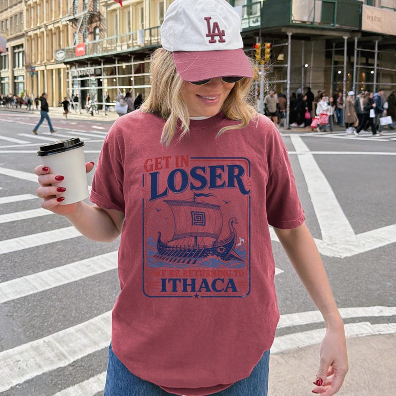Puede incluir: Una camiseta de manga corta color granate con un gr&aacute;fico azul de un barco vikingo y el texto GET IN LOSER WE'RE RETURNING TO ITHACA. La camiseta se combina con una gorra de b&eacute;isbol blanca y granate con las letras LA bordadas en el frente.