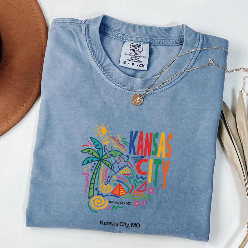 Peut inclure: Un t-shirt bleu &agrave; manches courtes avec un imprim&eacute; graphique repr&eacute;sentant un palmier, un joueur de football et le texte Kansas City 2026. Le t-shirt porte une &eacute;tiquette Comfort Colors indiquant la taille S et le texte Kansas City, MO.