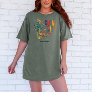 Peut inclure: Un t-shirt &agrave; manches courtes vert olive avec un imprim&eacute; graphique color&eacute; sur la poitrine. Le motif comprend un palmier, des formes abstraites et le texte KANSAS CITY en lettres grasses multicolores, avec Kansas City, MO imprim&eacute; en dessous.