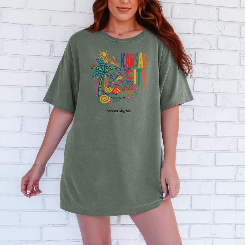 Peut inclure: Un t-shirt &agrave; manches courtes vert olive avec un imprim&eacute; graphique color&eacute; sur la poitrine. Le motif comprend un palmier, des formes abstraites et le texte KANSAS CITY en lettres grasses multicolores, avec Kansas City, MO imprim&eacute; en dessous.