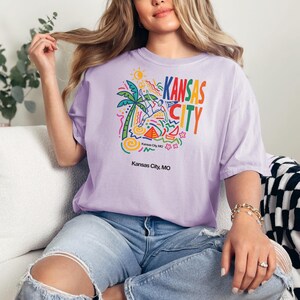 Peut inclure: Un t-shirt violet clair avec une impression graphique color&eacute;e portant le texte KANSAS CITY, un joueur de football stylis&eacute;, un palmier, un soleil et des formes g&eacute;om&eacute;triques. Le texte Kansas City, MO est imprim&eacute; en noir sous le motif principal.