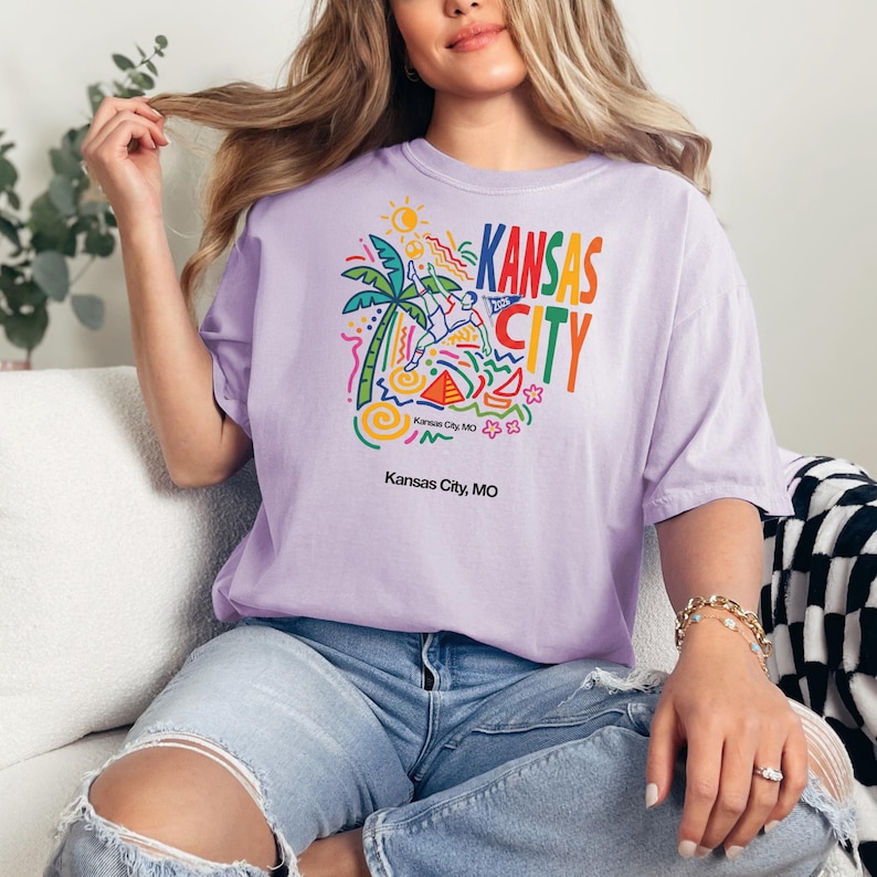 Peut inclure: Un t-shirt violet clair avec une impression graphique color&eacute;e portant le texte KANSAS CITY, un joueur de football stylis&eacute;, un palmier, un soleil et des formes g&eacute;om&eacute;triques. Le texte Kansas City, MO est imprim&eacute; en noir sous le motif principal.