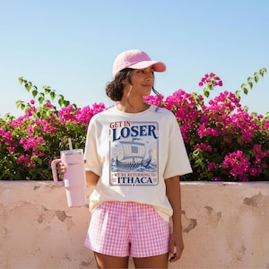 Puede incluir: Una camiseta de color claro con un gr&aacute;fico de un barco y el texto "GET IN LOSER WE'RE RETURNING TO ITHACA", combinada con pantalones cortos de cuadros vichy rosas, una gorra de b&eacute;isbol rosa claro y un vaso t&eacute;rmico rosa claro con pajita.