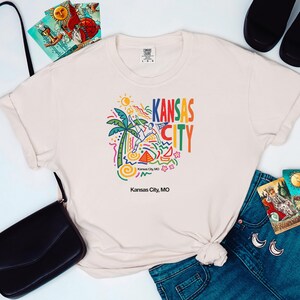 Peut inclure: Un t-shirt &agrave; manches courtes de couleur cr&egrave;me avec un imprim&eacute; graphique color&eacute; comprenant un palmier, un soleil, des formes abstraites et le texte Kansas City avec un fanion 2026. Le texte Kansas City, MO est imprim&eacute; sous le motif principal.