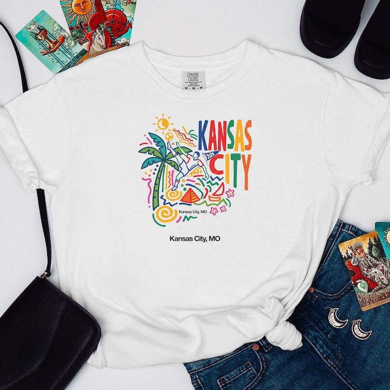 Peut inclure: Un t-shirt blanc &agrave; manches courtes avec un imprim&eacute; graphique color&eacute; portant le texte Kansas City. Le motif comprend un palmier, un soleil, un joueur de football et des formes abstraites en bleu, vert, jaune, orange et rose. Le texte Kansas City, MO est imprim&eacute; sous le motif principal.