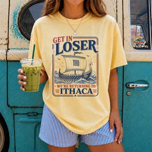Puede incluir: Una camiseta de manga corta de color amarillo p&aacute;lido con un gr&aacute;fico de un barco trirreme griego en el oc&eacute;ano. El texto dice GET IN LOSER WE'RE RETURNING TO ITHACA. El dise&ntilde;o est&aacute; enmarcado en un borde rectangular con detalles en azul y rojo.