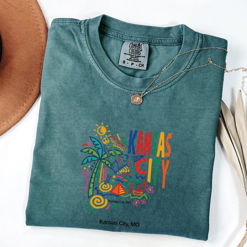 Peut inclure: Un t-shirt &agrave; manches courtes bleu sarcelle avec un col rond et une &eacute;tiquette Comfort Colors. L'avant pr&eacute;sente un graphique color&eacute; avec un palmier, un soleil, une pyramide et le texte KANSAS CITY en lettres grasses, avec Kansas City, MO imprim&eacute; en dessous.