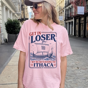 Puede incluir: Una camiseta de manga corta de color rosa claro con un estampado gr&aacute;fico en azul oscuro y rojo de un barco vikingo en el oc&eacute;ano. El texto dice GET IN LOSER WE'RE RETURNING TO ITHACA en una fuente estilizada, enmarcado por un dise&ntilde;o rectangular.