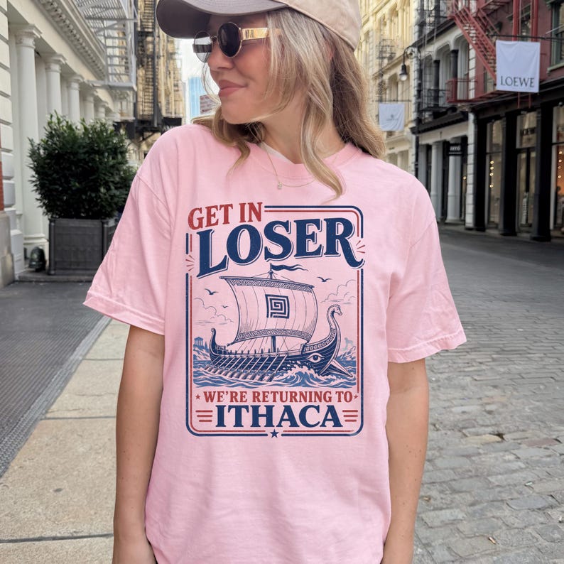 Puede incluir: Una camiseta de manga corta de color rosa claro con un estampado gr&aacute;fico en azul oscuro y rojo de un barco vikingo en el oc&eacute;ano. El texto dice GET IN LOSER WE'RE RETURNING TO ITHACA en una fuente estilizada, enmarcado por un dise&ntilde;o rectangular.