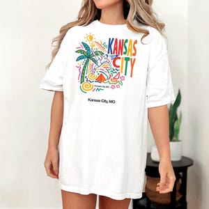 Peut inclure: Un t-shirt blanc &agrave; manches courtes avec un imprim&eacute; graphique color&eacute; comprenant le texte Kansas City, un palmier, un soleil et des formes abstraites. Sous le motif principal, le texte Kansas City, MO est imprim&eacute; en police noire simple.