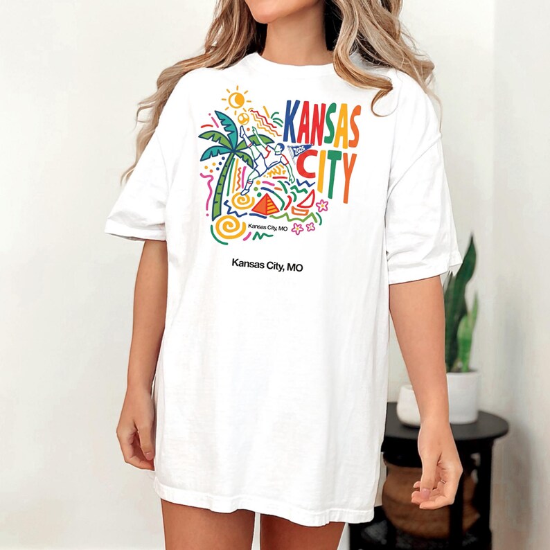 Peut inclure: Un t-shirt blanc &agrave; manches courtes avec un imprim&eacute; graphique color&eacute; comprenant le texte Kansas City, un palmier, un soleil et des formes abstraites. Sous le motif principal, le texte Kansas City, MO est imprim&eacute; en police noire simple.