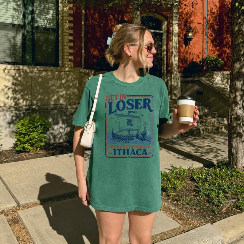 Puede incluir: Una camiseta de manga corta color verde bosque con un gr&aacute;fico de un barco trirreme griego y el texto "GET IN LOSER WE'RE RETURNING TO ITHACA" en azul y rojo. La camiseta se combina con un bolso de hombro blanco y un vaso de caf&eacute; desechable.