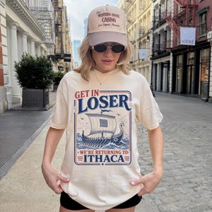 Puede incluir: Una camiseta color crema con un gr&aacute;fico de un barco vikingo en el oc&eacute;ano y el texto "GET IN LOSER WE'RE RETURNING TO ITHACA" en azul y rojo. La camiseta se combina con una gorra de b&eacute;isbol beige con el texto "THE WESTERN HOUSE CASINO".