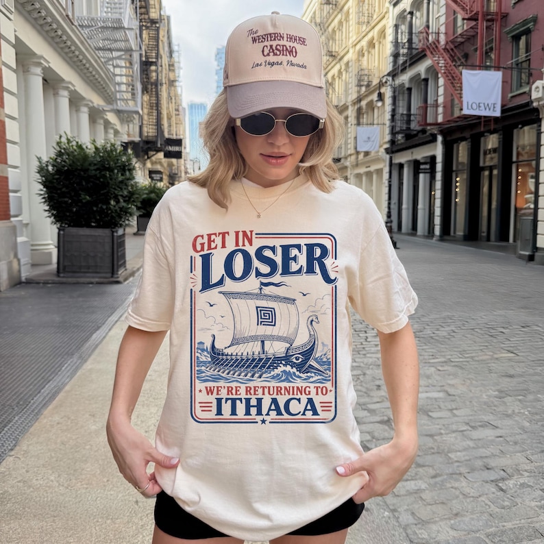 Puede incluir: Una camiseta color crema con un gr&aacute;fico de un barco vikingo en el oc&eacute;ano y el texto "GET IN LOSER WE'RE RETURNING TO ITHACA" en azul y rojo. La camiseta se combina con una gorra de b&eacute;isbol beige con el texto "THE WESTERN HOUSE CASINO".