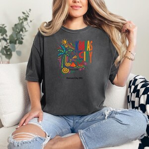 Peut inclure: Un t-shirt gris anthracite &agrave; manches courtes avec un imprim&eacute; graphique color&eacute; comprenant un palmier, un soleil et des formes abstraites. Le texte "KANSAS CITY" est imprim&eacute; en grandes lettres multicolores, avec "Kansas City, MO" en dessous.