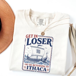 Puede incluir: Una camiseta de manga corta color blanco roto con un gr&aacute;fico de un barco vikingo en el oc&eacute;ano y el texto "GET IN LOSER WE'RE RETURNING TO ITHACA". La camiseta tiene una etiqueta de la marca Comfort Colors en el cuello y un collar dorado fino.