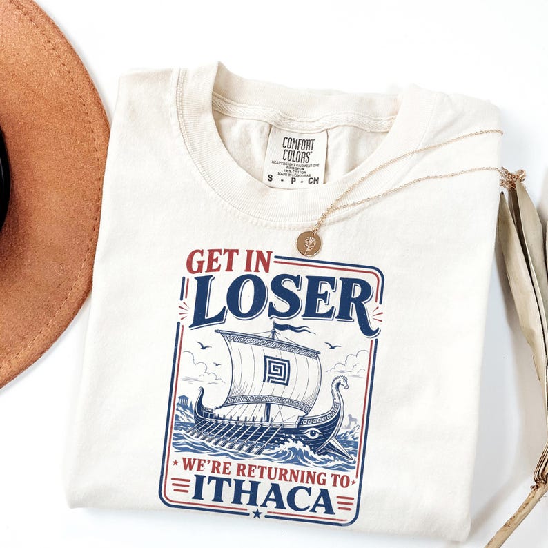 Puede incluir: Una camiseta de manga corta color blanco roto con un gr&aacute;fico de un barco vikingo en el oc&eacute;ano y el texto "GET IN LOSER WE'RE RETURNING TO ITHACA". La camiseta tiene una etiqueta de la marca Comfort Colors en el cuello y un collar dorado fino.