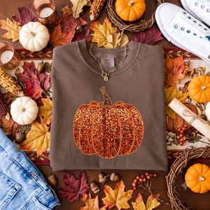 May include: A brown crewneck sweatshirt featuring a large, central graphic of a pumpkin with a mosaic-style orange and red pattern. A gold-toned chain necklace rests on the collar of the garment, which is displayed flat.