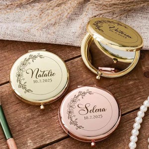 Może przedstawiać: Trzy spersonalizowane lusterka kompaktowe w kolorze złotym i różowego złota. Każde lusterko ma kwiatowy wzór i imię z datą: "Natalie 10.7.2025", "Victoria 10.7.2025" i "Selena 10.7.2025". Jedno lusterko jest otwarte, ukazując powierzchnię odbijającą światło.
