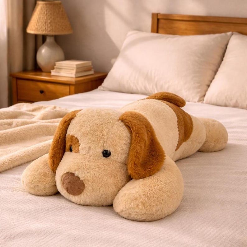 Puede incluir: Un gran peluche con forma de perro, en beige y marr&oacute;n, tumbado en una cama. El juguete tiene orejas ca&iacute;das, una mancha marr&oacute;n cerca de un ojo y una nariz marr&oacute;n. El perro est&aacute; sobre una colcha blanca con una manta beige.