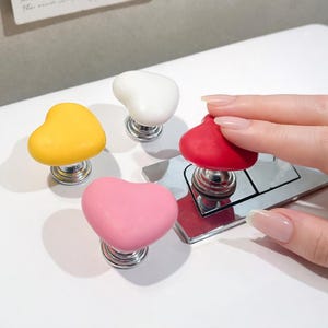 Op de afbeelding: Vier hartvormige kastknoppen in geel, wit, roze en rood. Elke knop heeft een zilveren basis en wordt weergegeven op een wit oppervlak. De knoppen staan in een rij, met een hand die de rode knop aanraakt.