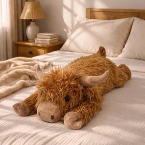 Puede incluir: Un peluche de vaca de las Tierras Altas marr&oacute;n tumbado en una cama. El juguete tiene un pelaje lanudo, cuernos curvados y un hocico suave. La vaca mide aproximadamente 76 cm de largo.