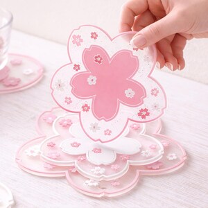 Può includere: Una pila di sottobicchieri a forma di fiore rosa e bianco. Il sottobicchiere superiore è tenuto in mano. I sottobicchieri hanno un design floreale con un centro rosa e piccoli accenti floreali. I sottobicchieri sono realizzati in materiale traslucido.