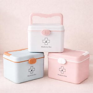 Könnte beinhalten: Drei Medikamentenboxen in Weiß, Hellblau und Rosa. Jede Box hat einen Griff, einen Verschluss und ein Kreuzsymbol mit den Worten "Medicine Box" darunter. Die Boxen sind übereinander gestapelt.