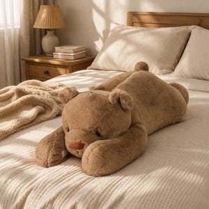 Puede incluir: Un gran oso de peluche marr&oacute;n claro tumbado en una cama con una colcha color crema. El oso tiene una cabeza redonda, orejas peque&ntilde;as y una nariz roja. La cama est&aacute; en una habitaci&oacute;n bien iluminada con una mesita de noche de madera y una l&aacute;mpara.