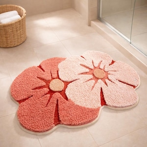 Peut inclure: Un tapis de bain en forme de fleur, de couleur corail et rose clair. Le tapis a une surface texturée et moelleuse, avec une bordure crème. Le motif présente deux grandes formes de fleurs avec un détail central circulaire. Le tapis est posé sur un sol carrelé.