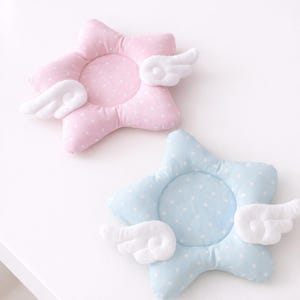 May include: Two star-shaped baby pillows, one pink and one blue, each with white polka dots and attached white wings. The pillows are designed for infants and are made of soft, plush material.