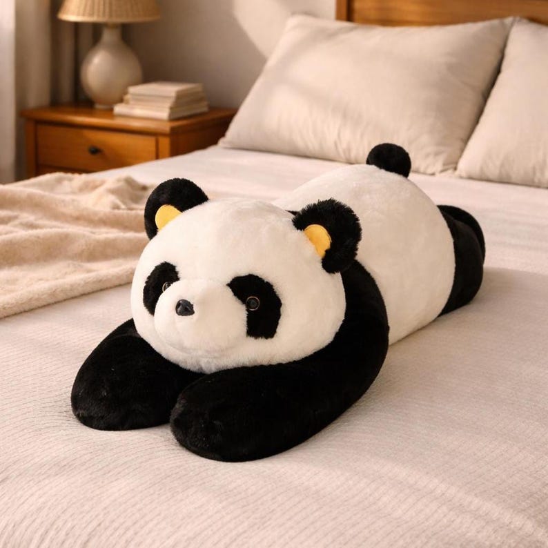 Puede incluir: Un gran coj&iacute;n de oso panda de peluche en posici&oacute;n de reposo. El panda tiene un cuerpo blanco con brazos, piernas y orejas negras. La cara del panda tiene parches negros en los ojos y una peque&ntilde;a nariz negra. Las orejas tienen detalles amarillos.