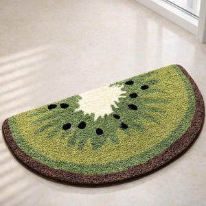 May include: A half-circle rug designed to resemble a kiwi fruit slice. The rug features shades of green, a white center, and black seed details. A brown border frames the design. The rug is made of a soft, textured material.
