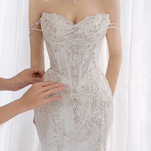 May include: A white strapless gown featuring an intricate bodice with a sweetheart neckline, adorned with extensive silver beadwork, clear crystals, and delicate draped pearl-like strands. The garment includes a matching high-neck choker collar.