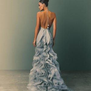 Puede incluir: Un vestido de fiesta azul claro sin espalda con una falda con volantes en cascada. El vestido presenta una delicada correa halter con cuentas y una cola fluida. El cabello de la modelo está recogido en un moño. El fondo es verde apagado.