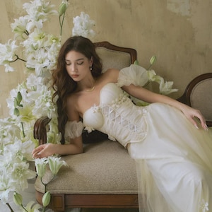 Puede incluir: Un vestido color crema con hombros descubiertos, corpiño estilo corsé y falda fluida. El vestido presenta adornos florales y lo lleva una persona reclinada en una silla vintage. Lirios blancos rodean a la persona y la silla.