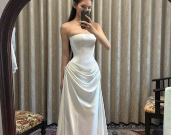 May include: A floor-length white strapless gown featuring a fitted bodice and a draped, gathered skirt detail at the waist. The dress is made of a smooth, solid white fabric that flows down to the floor in a clean, simple, and elegant silhouette.