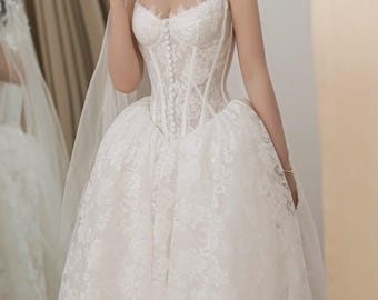 May include: A white strapless wedding dress featuring a corset-style bodice with a vertical row of small buttons and a sweetheart neckline with delicate lace trim. The full skirt is made of floral lace fabric, and a sheer veil is draped over the shoulders.