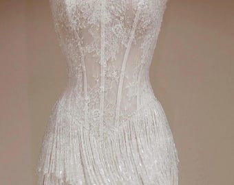 May include: A white strapless mini dress featuring a corset-style bodice with intricate lace overlays and vertical boning. The skirt is adorned with multiple layers of long, shimmering white fringe. A matching lace choker is included.