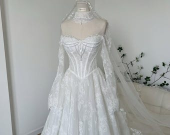 Pode incluir: Um vestido de noiva branco longo até ao chão com um corpete estilo espartilho com detalhes em pérolas, mangas compridas de renda transparente e gola alta de renda. O vestido inclui um véu de renda a condizer sobre os ombros.