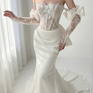 Könnte beinhalten: Ein weißes Brautkleid mit trägerlosem Oberteil und fließendem Rock. Das Oberteil hat aufwendige Spitzendetails und florale Verzierungen. Die langen Ärmel sind aus Spitze und mit Schleifen versehen.