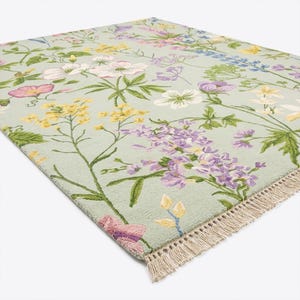 Puede incluir: Una alfombra rectangular con un estampado floral. La alfombra presenta un fondo verde claro con flores bordadas en tonos de rosa, amarillo, morado y blanco. La alfombra tiene un borde con flecos.