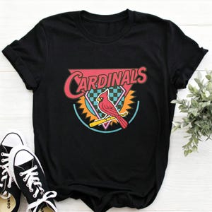 May include: A black short-sleeve t-shirt featuring a graphic print of a red cardinal bird perched on a yellow baseball bat. The design includes the word CARDINALS in pink, set against a background of a pink triangle, a teal circle, and a checkered pattern.