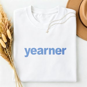 Può includere: Una t-shirt bianca a maniche corte con la parola yearner stampata in un carattere sans-serif azzurro al centro del petto. Una delicata collana a catena color argento con un piccolo ciondolo a forma di stella poggia sullo scollo.