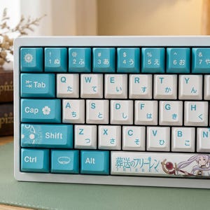 Op de afbeelding: Een witte mechanische toetsenbord met lichtblauwe en witte keycaps. De keycaps bevatten Japanse tekens en Engelse letters. Het toetsenbord heeft een aangepast ontwerp met de tekst "葬送のフリーレン" en een anime-personage-illustratie.