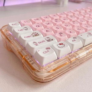 Puede incluir: Un teclado mecánico rosa y blanco con teclas personalizadas. El teclado tiene una carcasa transparente teñida de rosa. Las teclas son rosas y blancas, con ilustraciones personalizadas de un gato de dibujos animados. El teclado está diseñado para uso informático.