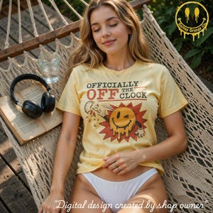 Może przedstawiać: Jasnożółty t-shirt z krótkim rękawem z grafiką topniejącej, puszczającej oko buźki oraz napisem "OFFICIALLY OFF THE CLOCK" w ciemnoczerwonych i czarnych literach. Projekt zawiera małą ikonę zegara i czerwone tło w kształcie gwiazdy.
